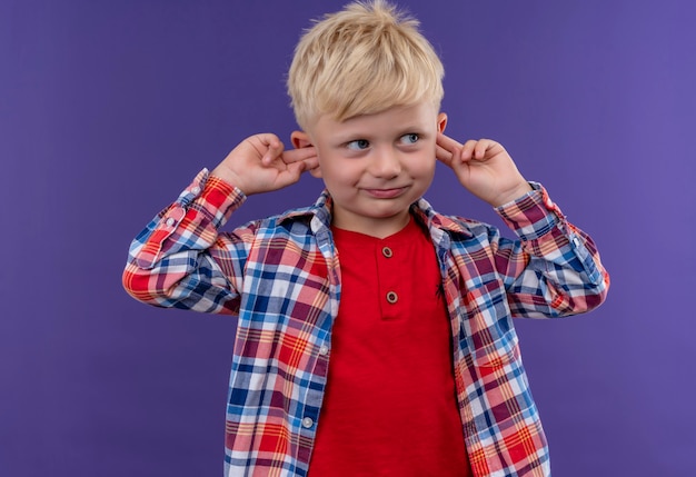 Foto grátis um garotinho fofo com cabelo loiro vestindo uma camisa xadrez segurando os dedos nas orelhas em uma parede roxa