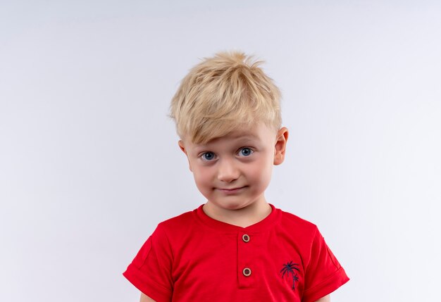 Um garotinho lindo com cabelo loiro e olhos azuis vestindo uma camiseta vermelha parecendo surpreendentemente em uma parede branca