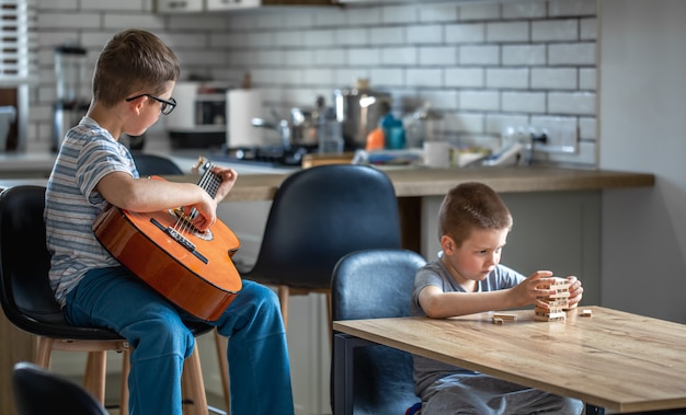 Um garotinho toca violão, e seu irmão constrói uma torre com cubos de madeira em casa na mesa.