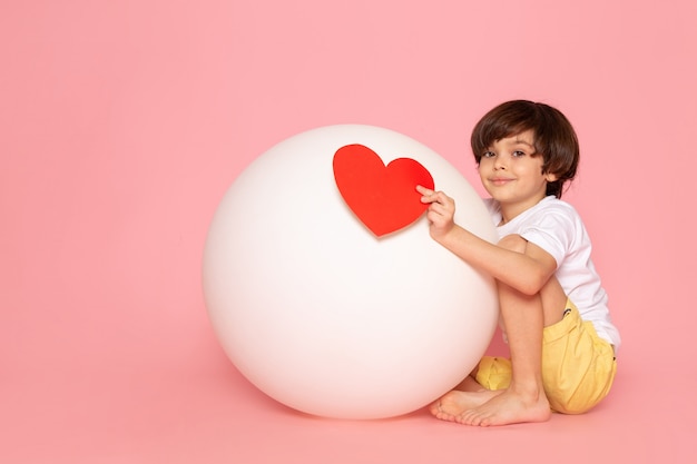Um garoto engraçado bonito vista frontal em camiseta branca segurando coração forma brincando com bola branca redonda no chão rosa