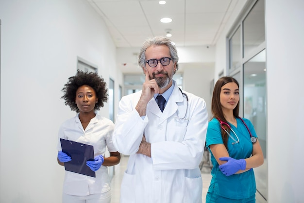 Um grupo multiétnico de três médicos e enfermeiros em um corredor de hospital vestindo jalecos e casacos a equipe de profissionais de saúde está olhando para a câmera e sorrindo