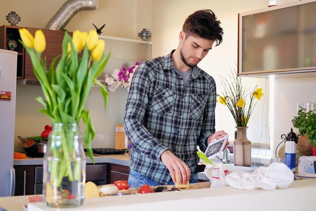 Um homem barbudo bonito vestido com uma camisa de lã fazendo salada de legumes na cozinha de casa.