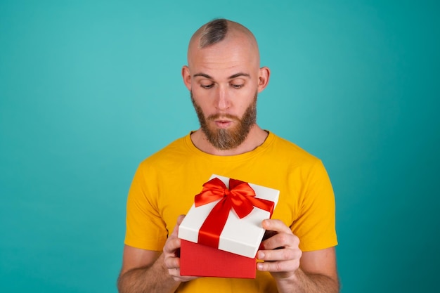 Um homem barbudo com uma camiseta laranja em uma parede turquesa com uma caixa de presente em um humor exultante, dá alegria, sorri agradavelmente