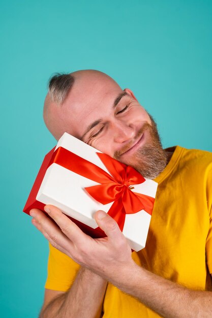 Um homem barbudo com uma camiseta laranja em uma parede turquesa com uma caixa de presente em um humor exultante, dá alegria, sorri agradavelmente