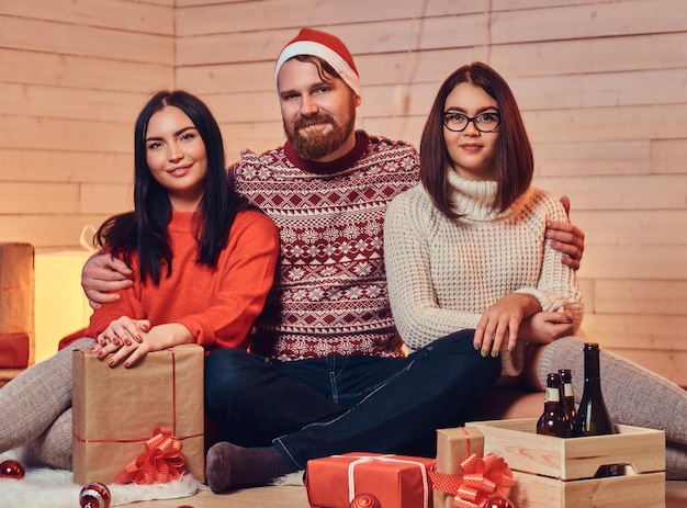 Um homem barbudo e duas mulheres morenas festa de Natal sobre parede de madeira branca.