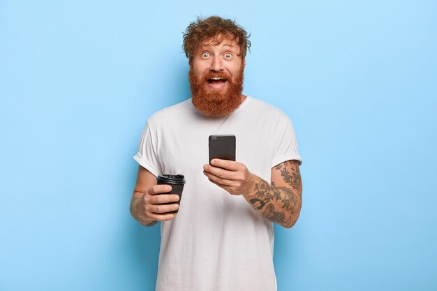 Um homem barbudo e emotivo sorridente tem cabelo ruivo, segura um telefone celular, compartilha ótimas notícias com um amigo, olha com um sorriso largo e olhos esbugalhados, usa uma camiseta branca casual, segura um café para viagem