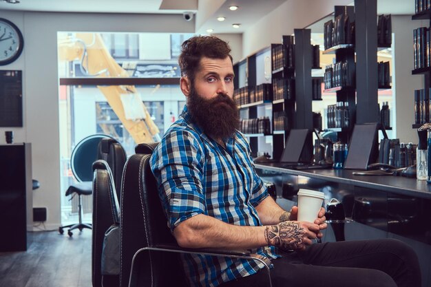 Um homem barbudo elegante bonito com uma tatuagem no braço vestido com uma camisa de flanela bebe café em uma barbearia.