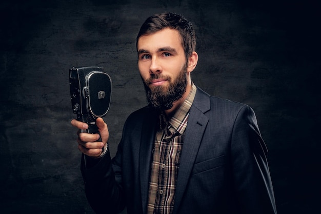 Um homem barbudo elegante, vestido de terno, segura uma câmera de vídeo vintage de 8 mm.
