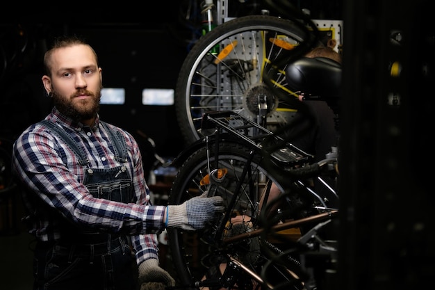 Foto grátis um homem bonito e elegante vestindo uma camisa de flanela e macacão jeans, trabalhando com uma roda de bicicleta em uma oficina. um trabalhador usando uma chave monta a roda em uma bicicleta em uma oficina.