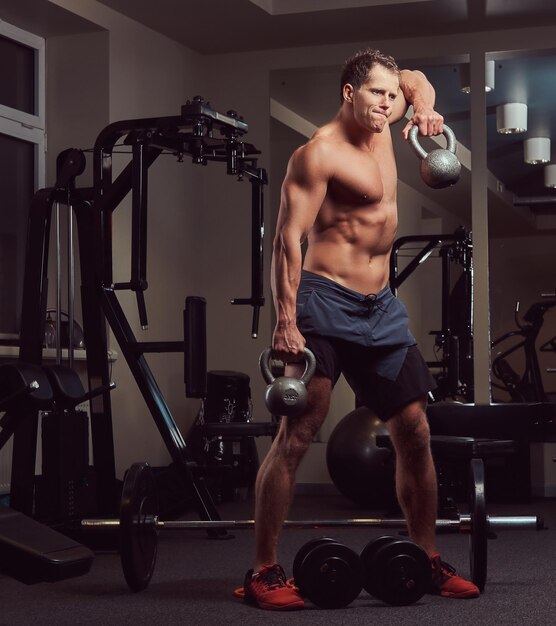 Um homem bonito musculoso fisiculturista sem camisa fazendo exercícios nos músculos deltóides com um kettlebell no ginásio.