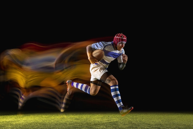 Um homem caucasiano jogando rugby no estádio em luz mista. Ajuste o jovem jogador do sexo masculino em movimento ou ação durante o jogo de esporte. Conceito de movimento, esporte, estilo de vida saudável.