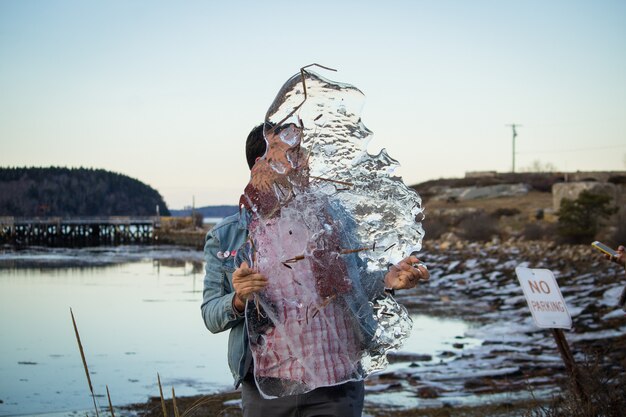 Um homem caucasiano, segurando um pedaço gigantesco de gelo nas mãos com um lago no fundo