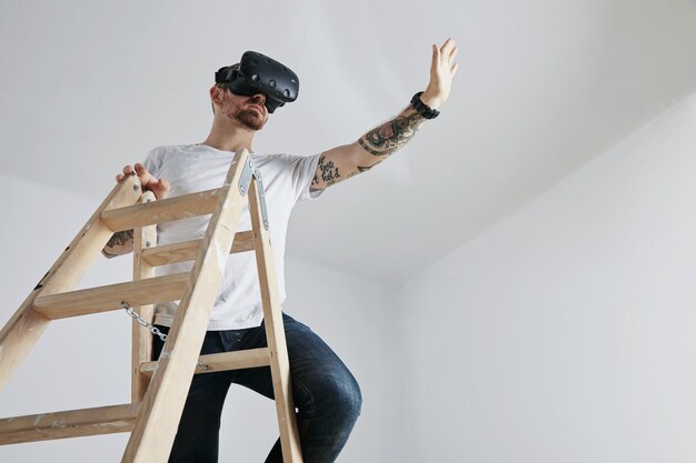 Um homem com tatuagens, vestindo uma camiseta branca lisa e óculos de realidade virtual no topo de uma escada de madeira jogando um jogo de realidade virtual