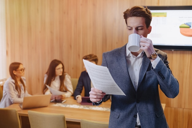 Um homem elegante de jaqueta e camisa com uma xícara de café na mão fica e lê documentos