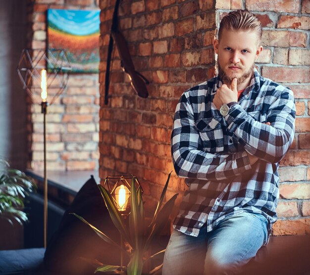 Foto grátis um homem loiro barbudo hipster vestido com jeans e camisa de lã posando em uma sala com interior loft.