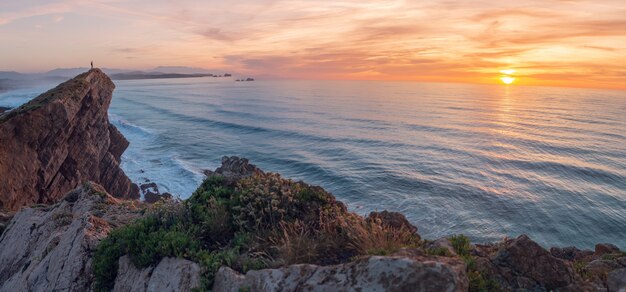 Um homem olha para o mar de um penhasco ao pôr do sol.