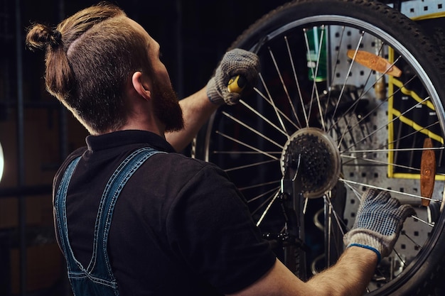 Um homem ruivo bonito em um macacão jeans, trabalhando com uma roda de bicicleta em uma oficina. Um trabalhador remove o pneu da bicicleta em uma oficina.