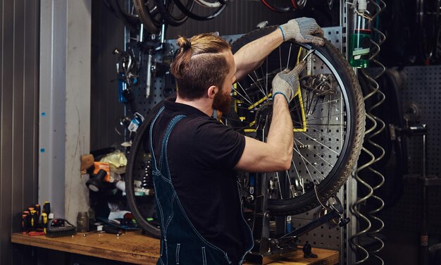 Um homem ruivo bonito em um macacão jeans, trabalhando com uma roda de bicicleta em uma oficina. Um trabalhador remove o pneu da bicicleta em uma oficina.