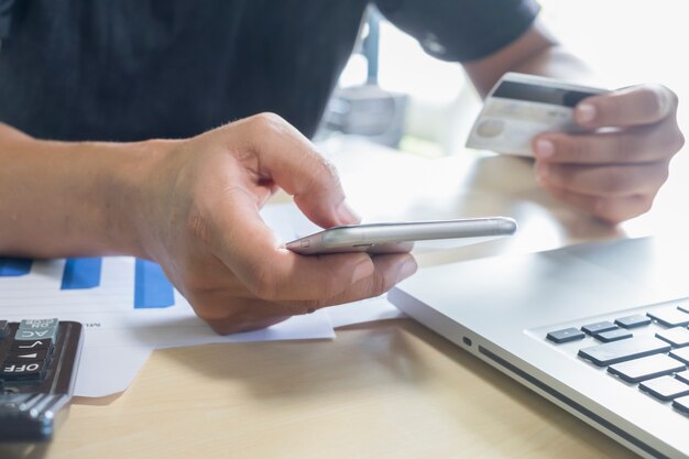 Um homem segurando telefone celular e cartão de crédito no laptop para compras on-line