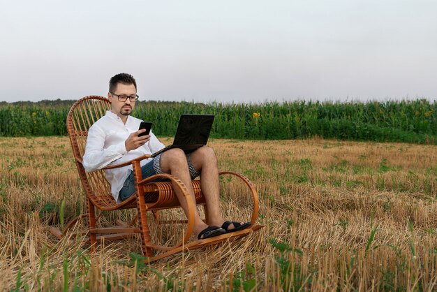 Um homem segurando um smartphone enquanto trabalha em um laptop sentado em uma cadeira de balanço no meio de um campo | Foto Premium