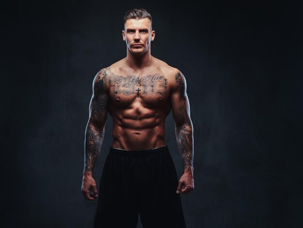 Um homem sem camisa musculoso tatuado com cabelo elegante posando para a câmera em um fundo escuro.