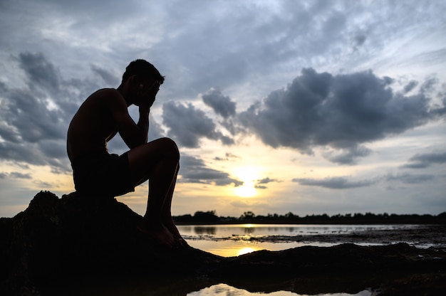 Um homem sentado dobrou os joelhos, segurando as mãos no rosto, na base da árvore, e havia água ao redor.