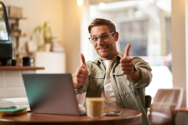 Foto grátis um homem sorridente e alegre com óculos, um homem com os polegares levantados, senta-se num café com um portátil e recomenda um site.