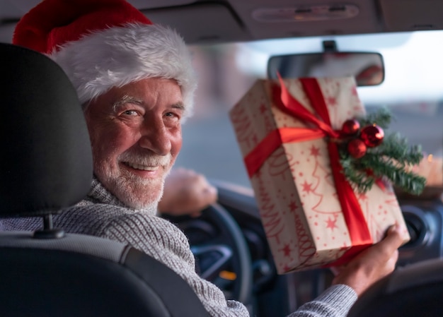 Um homem sorridente sênior dentro de seu carro com um chapéu de papai noel pronto para entrega em domicílio de presentes de natal. o velho avô barbudo com um grande sorriso