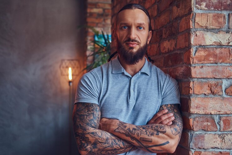 Foto grátis um homem tatuado bonito com um corte de cabelo elegante e barba, em uma camiseta cinza, encostado em uma parede de tijolos em uma sala com um interior loft.