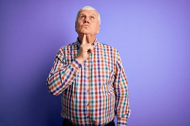 Um homem velho, bonito e cinzento, vestindo uma camisa colorida casual sobre um fundo roxo isolado, pensando concentrado na dúvida, com o dedo no queixo e olhando para cima, perguntando: