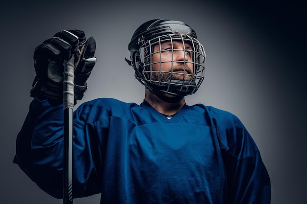 Um jogador de hóquei no gelo barbudo brutal no capacete de segurança segura o bastão de jogo no fundo cinza da vinheta.
