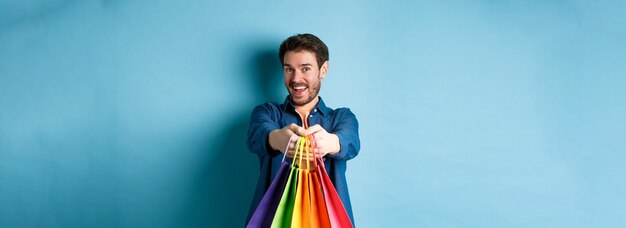 Um jovem alegre estende as mãos com sacolas de compras e sorri dando-lhe presentes em pé no azul