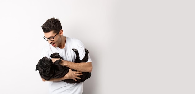 Um jovem bonito brincando com o dono de um cachorrinho preto fofo acariciando um pug em pé sobre fundo branco