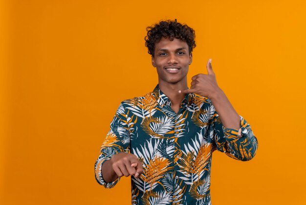 Um jovem bonito de pele escura com cabelo encaracolado e camisa estampada de folhas sorrindo mostrando gesto de me ligar