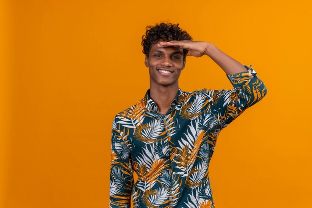 Um jovem bonito de pele escura com cabelo encaracolado em camisa estampada de folhas sorrindo e com as mãos na cabeça