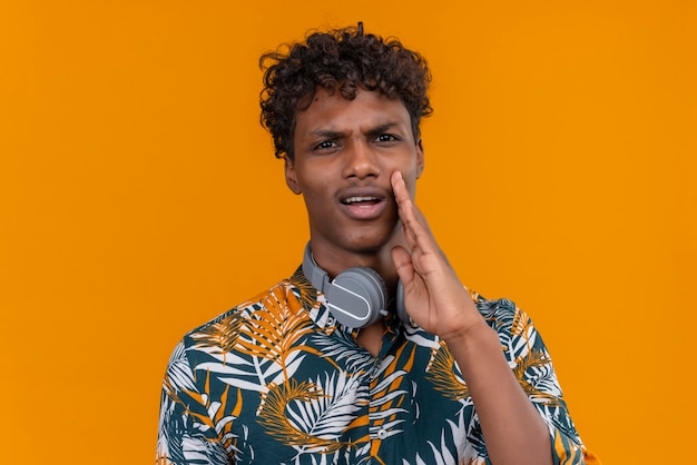 Um jovem bonito de pele escura com cabelo encaracolado em uma camisa estampada de folhas com rosto agressivo chamando alguém de mão no rosto