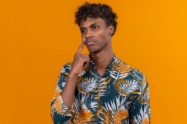 Um jovem bonito de pele escura com cabelo encaracolado em uma camisa estampada de folhas pensando com o dedo indicador no rosto