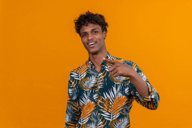 Um jovem bonito de pele escura com cabelo encaracolado em uma camisa estampada de folhas sorrindo e apontando com o dedo indicador