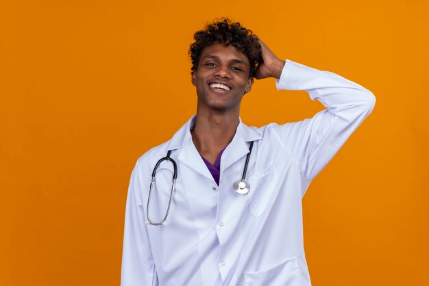 Um jovem bonito de pele escura com cabelo encaracolado, jaleco branco e estetoscópio segurando a mão na cabeça