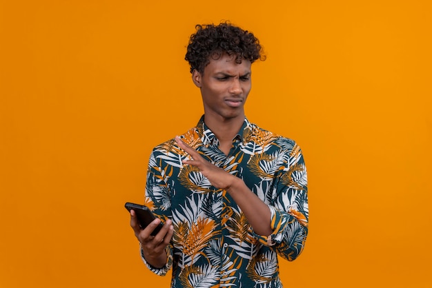 Um jovem bonito de pele escura com cabelo encaracolado numa camisa estampada de folhas com expressão de raiva segurando um telefone celular