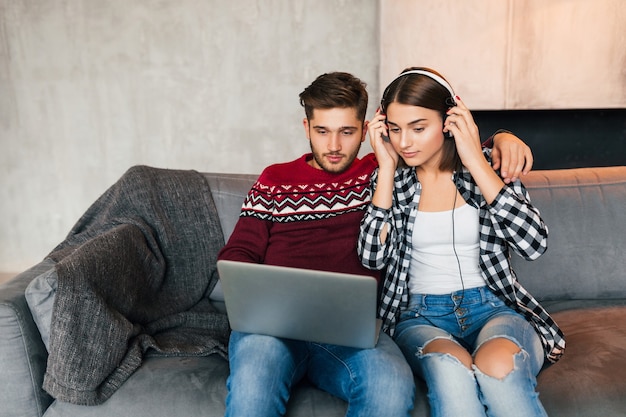 Um jovem bonito e uma mulher sentada em casa no inverno, olhando no laptop, ouvindo fones de ouvido, alunos estudando online, casais em momentos de lazer juntos,