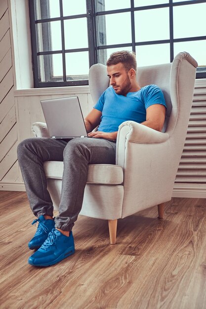 Um jovem bonito em uma camiseta azul e tênis trabalhando em seu laptop enquanto está sentado em uma cadeira.