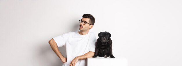 Foto grátis um jovem bonito parado perto de um pug preto fofo olhando bem com expressão arrogante em pé