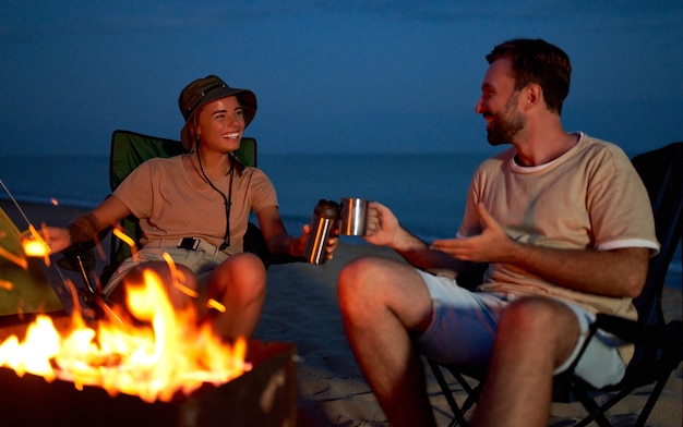 Um jovem casal apaixonado está sentado perto da tenda em cadeiras dobráveis perto do fogo, bebendo chá à noite na praia.