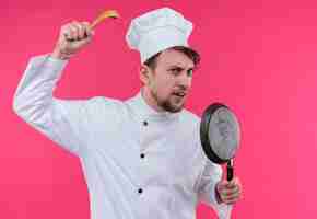 Foto grátis um jovem chef barbudo agressivo de uniforme branco segurando uma colher de pau com uma frigideira enquanto olha para uma parede rosa