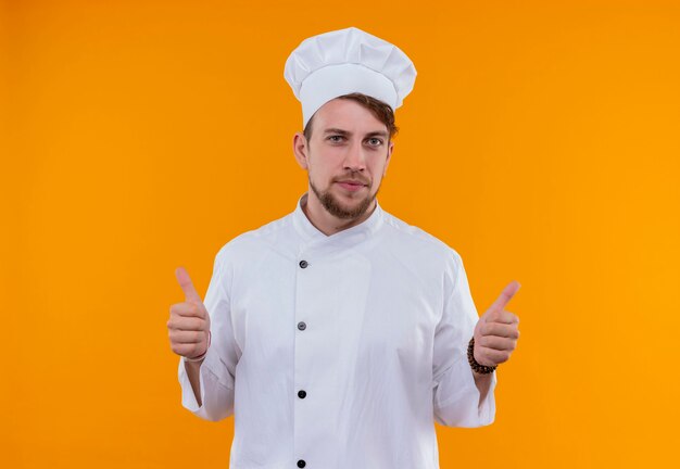 Um jovem chef barbudo com uniforme branco mostrando os polegares para cima enquanto olha para uma parede laranja