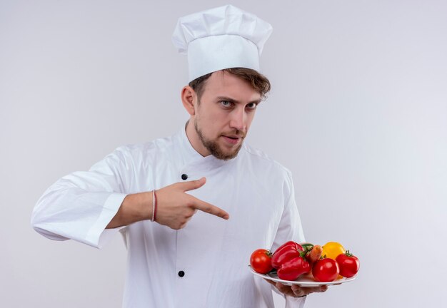 Um jovem chef barbudo confiante vestindo uniforme de fogão branco e chapéu apontando para um prato branco com legumes frescos em uma parede branca