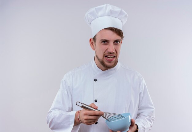 Um jovem chef barbudo descontente vestindo uniforme de fogão branco e chapéu segurando uma tigela azul com uma colher de batedeira enquanto olha para uma parede branca