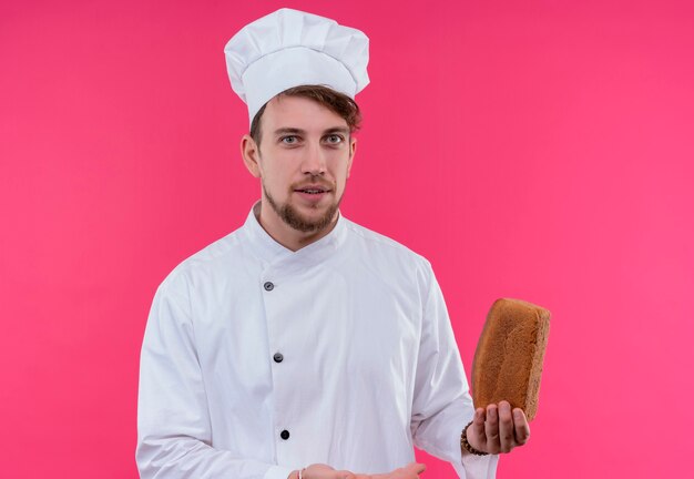 Um jovem chef barbudo e alegre, de uniforme branco, segurando um pedaço de pão enquanto olha para uma parede rosa