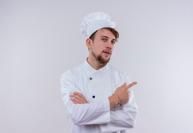 Um jovem chef barbudo e confiante vestindo uniforme branco de fogão e chapéu apontando para cima enquanto olha para uma parede branca